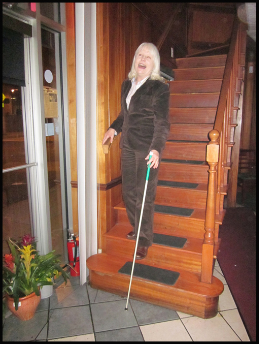two photos show Susan coming down the steps with her right hand on the end of the railing and left hand holding her white cane.  She has one foot on the third step from the bottom and is extending her other foot to the next stair, grinning as she opens her mouth as if screaming.